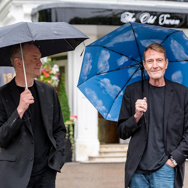 An image of two men outside The Old Swan hotel holding umbrellas and smiling.