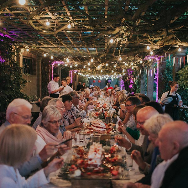 A giant table outside in a whimsical garden full of people enjoying a meal.