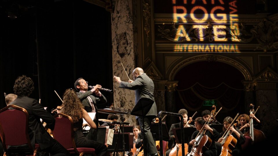 Chineke Orchestra take the stage with the conductor waving his baton, guiding a clarinetist, surrounded by the strings section.