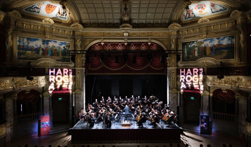 The curtain has risen for an orchestra playing at the Royal Hall in the centre of the image. There are elegant golden walls with classical paintings hung high up on them. Projected on the walls is the Harrogate International Festivals logo.