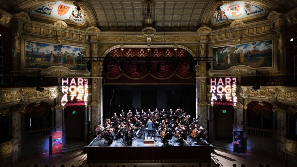 The curtain has risen for an orchestra playing at the Royal Hall in the centre of the image. There are elegant golden walls with classical paintings hung high up on them. Projected on the walls is the Harrogate International Festivals logo.