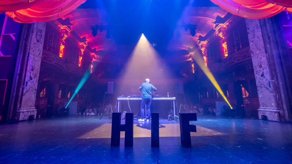 A person DJing on the HIF main stage. There are multi-coloured strobe lights projecting onto the crowd.