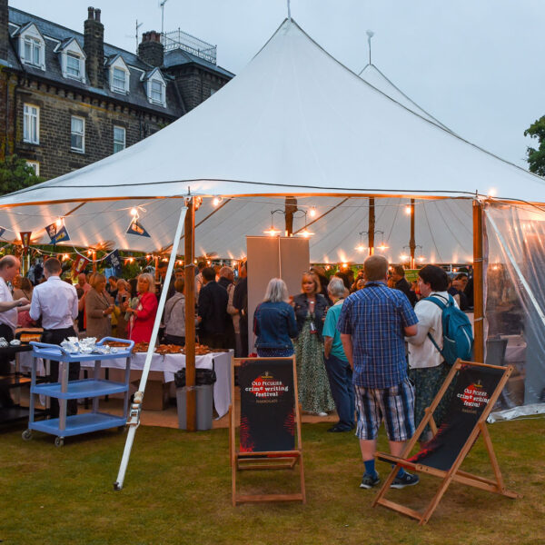 A marquee outside lit up with fairy lights. It is full of people inside and people stood outside looking in.