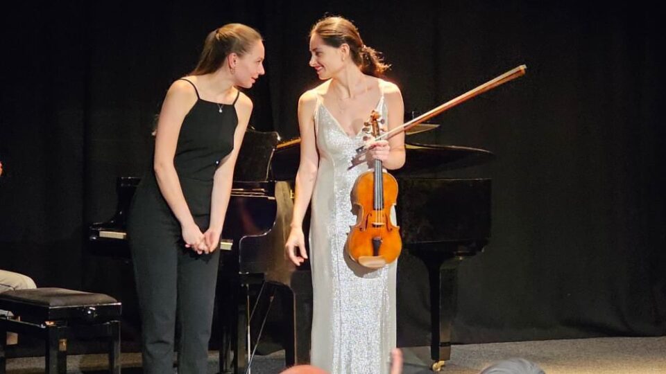 Violin player Esther Abrami on stage holding her cello and smiling at a person to her left.
