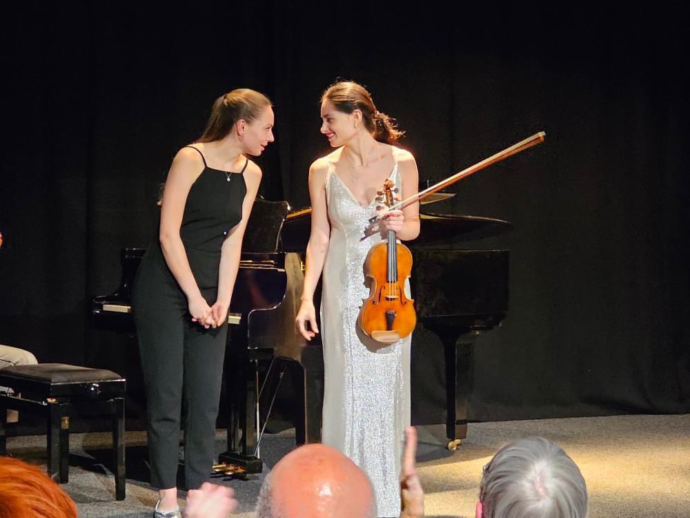Violin player Esther Abrami on stage holding her cello and smiling at a person to her left.