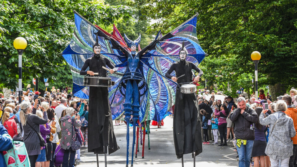 Three street performers on stilts holding snare drums. The performer in the middle of the two drummers is wearing a fantasy themed outfit featuring wings that come out of the arm sleeves.