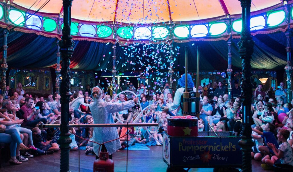 Some clowns onstage in the Spiegeltent. There is full audience who are laughing and there is lots of confetti in the air.
