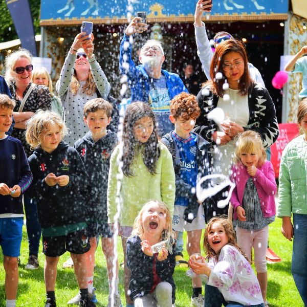 Children stood outside enjoying a water fountain spraying upwards whilst their parents take videos and photos on their phones of the action.