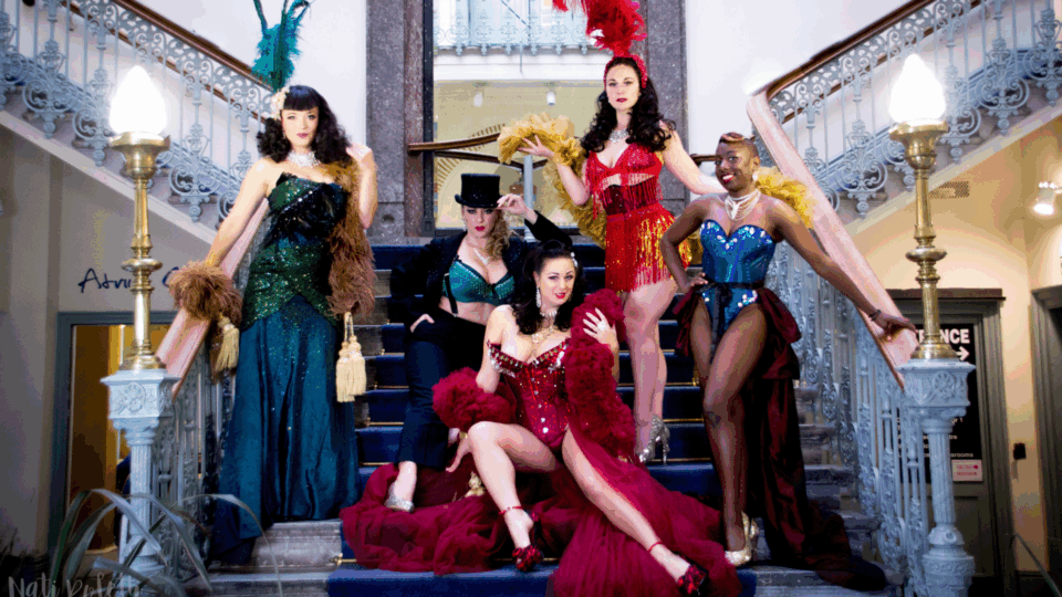 Five performers pose on a grand staircase inside an ornate building. They wear colourful, sequinned costumes with feathers and accessories, including one performer in a top hat and tuxedo‑style outfit.
