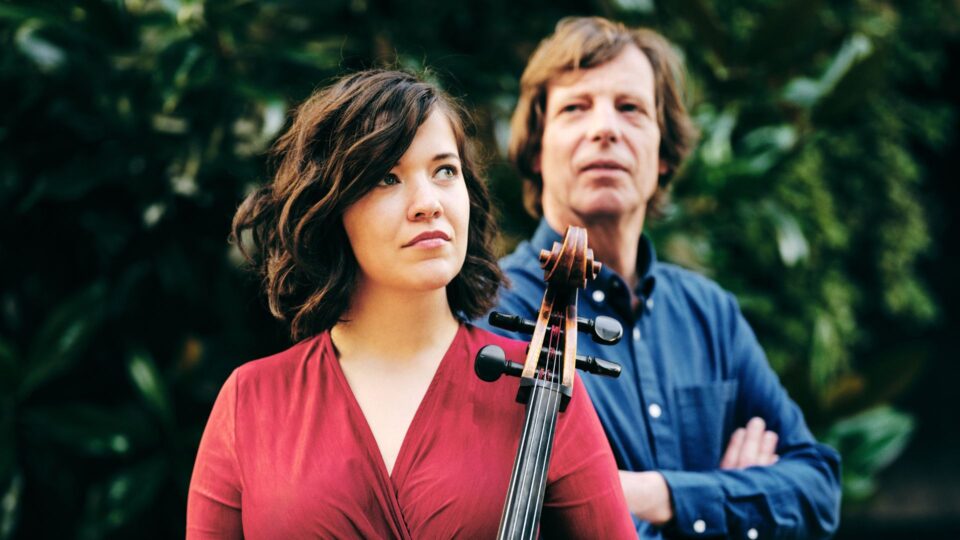A lady in a red dress is standing in front of a pianist in a blue shirt holding her cello.