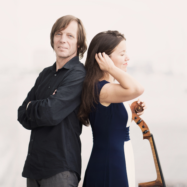 A pianist and a cellist are standing with their backs to each other. The lady is holding her cello.