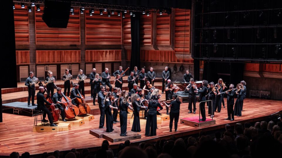 Full orchestra performing on a wooden stage with strings, woodwinds, and brass players, lit by warm stage lighting, with the audience visible in the foreground.