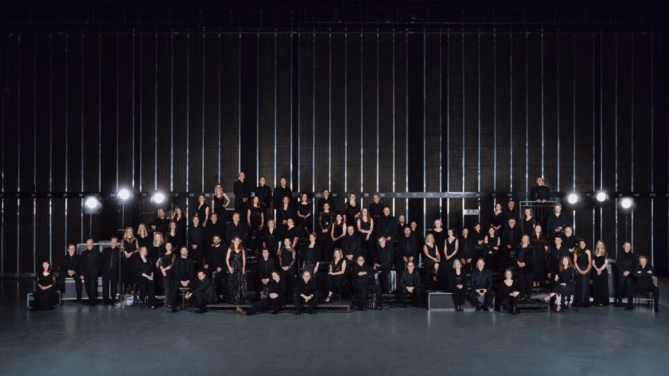 A large group of people dressed in black pose on tiered risers in a dark studio, facing the camera.