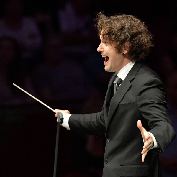 Conductor, Nicholas Collon, holding a baton and leading an ensemble on a darkened stage during a live concert.