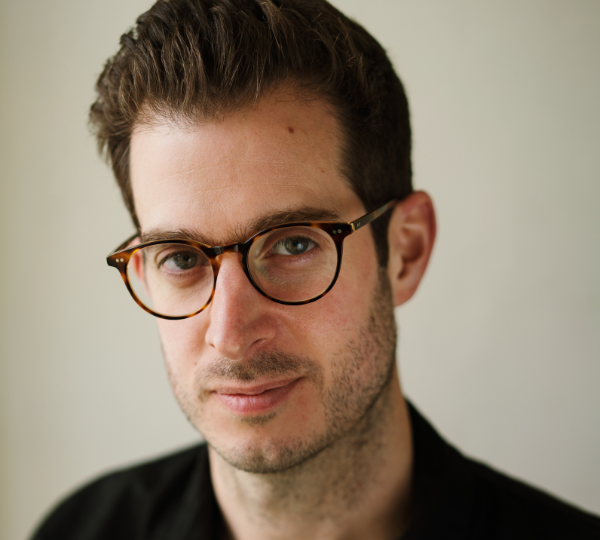 Dave Green, shown in a close-up portrait. He has short brown hair, round tortoiseshell glasses, and wears a dark shirt. He faces the camera against a plain light background.