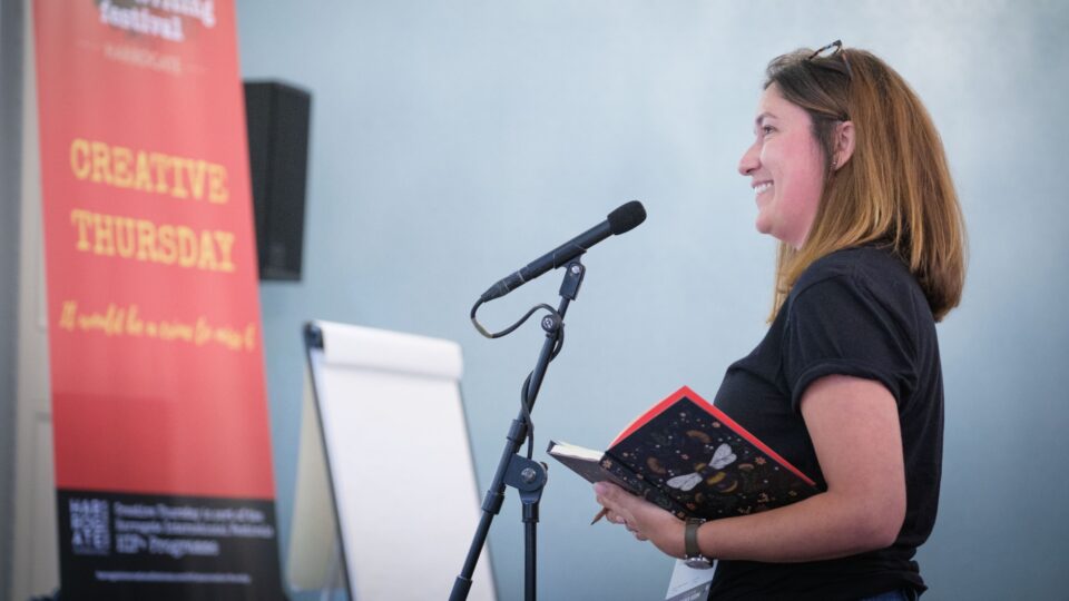 A woman speaks into a microphone while holding a notebook, standing beside a red event banner and a blank flip chart.