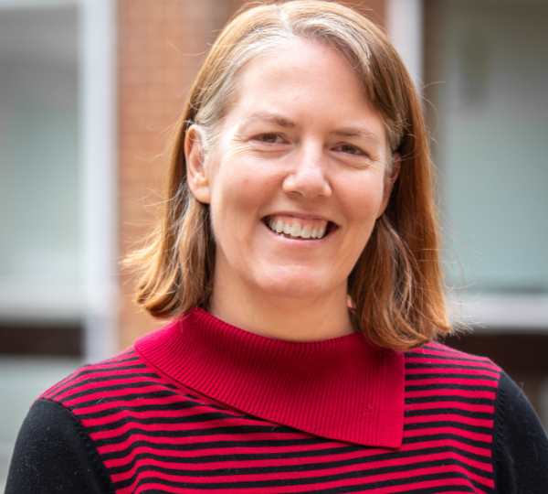 Dr Gillian Sandstrom outdoors in front of a brick and glass building. She has shoulder‑length light brown hair and wears a black top with red stripes and a red collar.