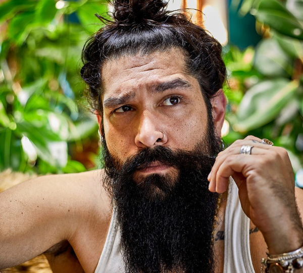 Johnny Balchandani in front of lush green plants. He has a long black beard, a top‑bun hairstyle, and wears a white tank top.