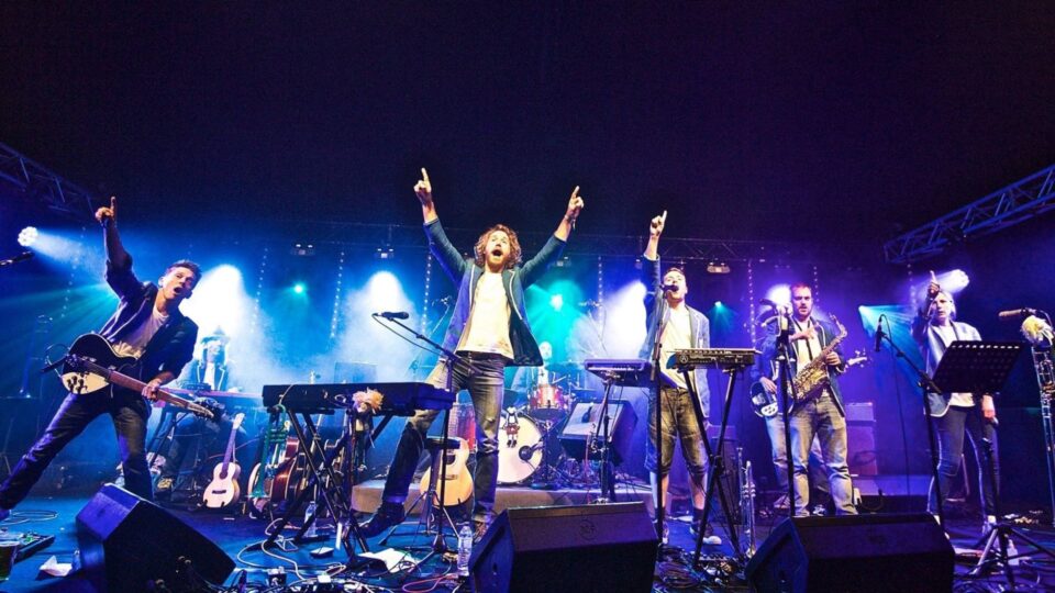 A live band performs on a brightly lit stage, with six musicians playing guitars, keyboards, saxophone, and drums.