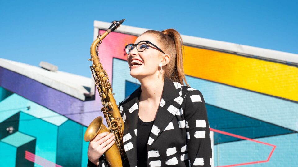 A person stands outdoors holding a saxophone in front of a brightly coloured geometric mural painted on a brick wall.