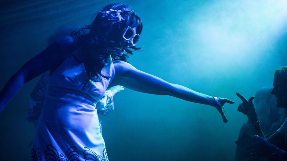 A performer in a white dress with patterned detailing, large white sunglasses, and a flower in their hair reaches an arm toward another person in a dim, blue‑lit setting.