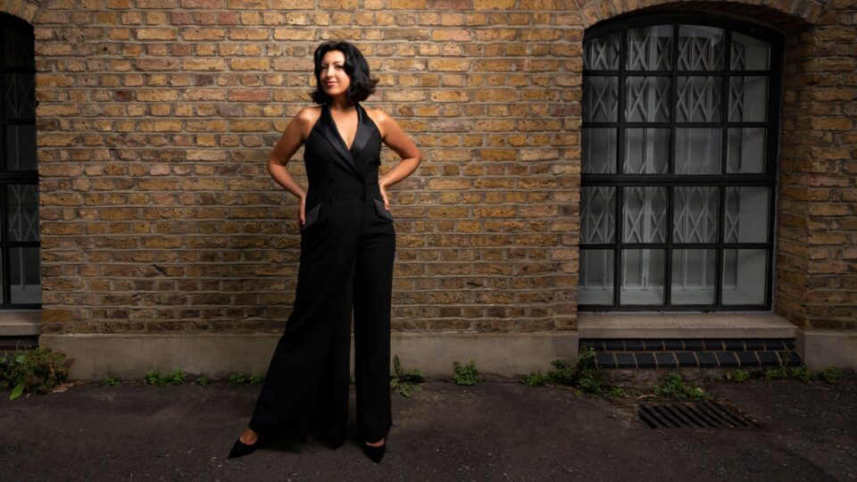 Alexandra stands outdoors in front of a brick wall with two arched windows. She has short dark hair and wears a sleeveless black jumpsuit with a deep V‑neck and black heels, standing with hands in pockets.