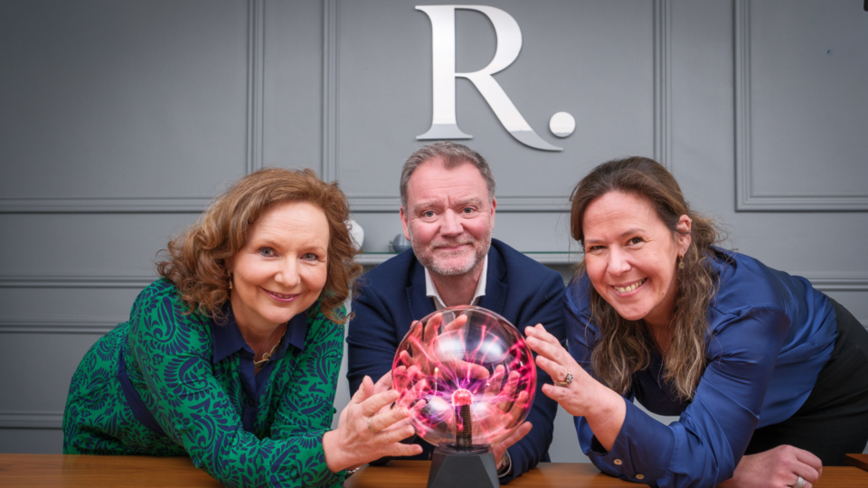 Three people—Rachel Tunnicliffe (left), Simon Morris (centre) and Sharon Canavar (right)—lean over a wooden table, smiling as they touch a glowing plasma ball.