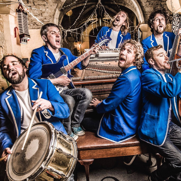 Six musicians wearing matching bright blue jackets perform together in a rustic indoor space with stone walls and string lights