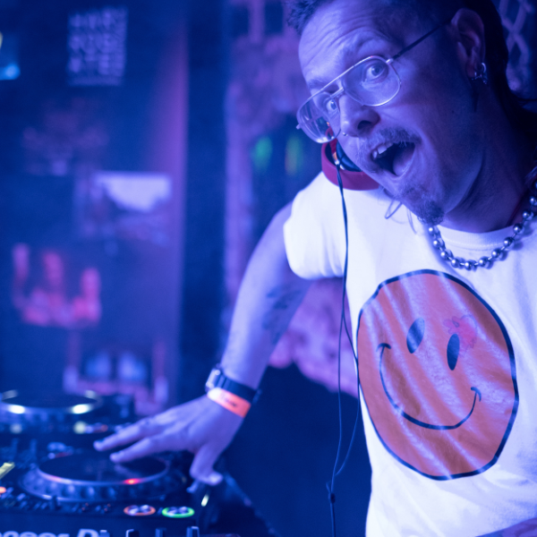 A person stands at a DJ booth operating turntables and a mixer, wearing a white T‑shirt with a large orange smiley face, glasses, a chain necklace, and headphones around their neck. Blue and purple lighting creates a nightclub‑style atmosphere.