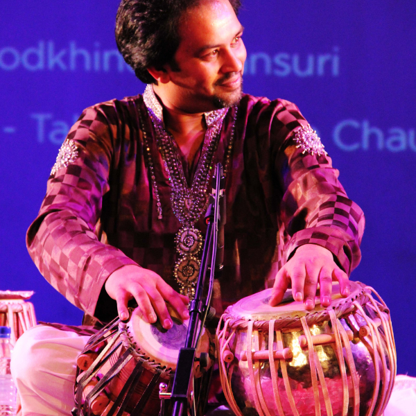 A musician in an ornate embroidered outfit plays a pair of tabla drums onstage, hands poised mid‑performance against a purple‑lit backdrop with partially visible projected text.