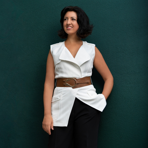Alexandra Dariescu stands against a dark teal wall wearing a sleeveless white wrap‑style top with a wide brown belt and black trousers. Their hands rest in the belt area.