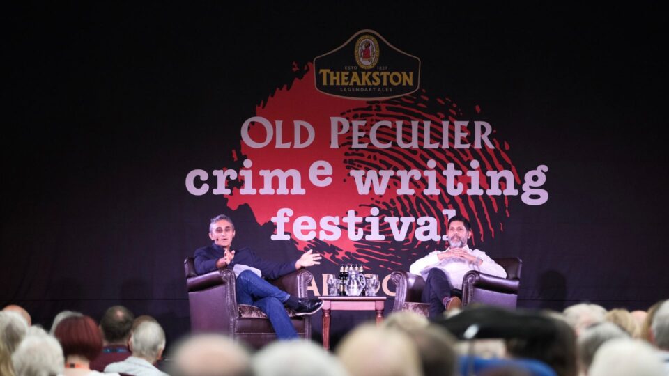 Two speakers sit in armchairs onstage at the Old Peculier Crime Writing Festival, talking in front of an audience. A large backdrop behind them shows the festival’s red fingerprint graphic and Theakston branding.