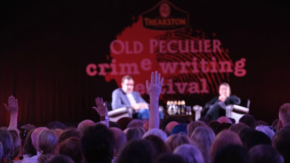 Two speakers sit onstage at the Old Peculier Crime Writing Festival while audience members raise their hands to ask questions. A large backdrop behind the stage displays the festival’s branding.