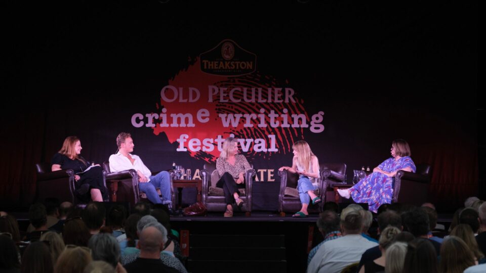 A panel discussion takes place onstage at the Theakston Old Peculier Crime Writing Festival. Five people sit facing the audience. The audience sits closely packed, watching the speakers