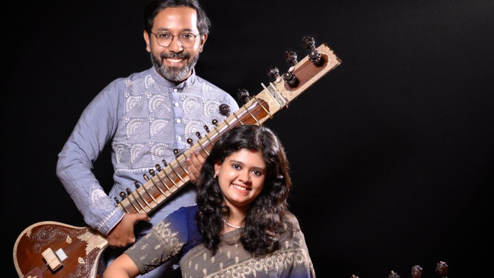 A standing musician holds a sitar while another person sits in front wearing an embroidered saree and smiling. Both pose against a dark background.
