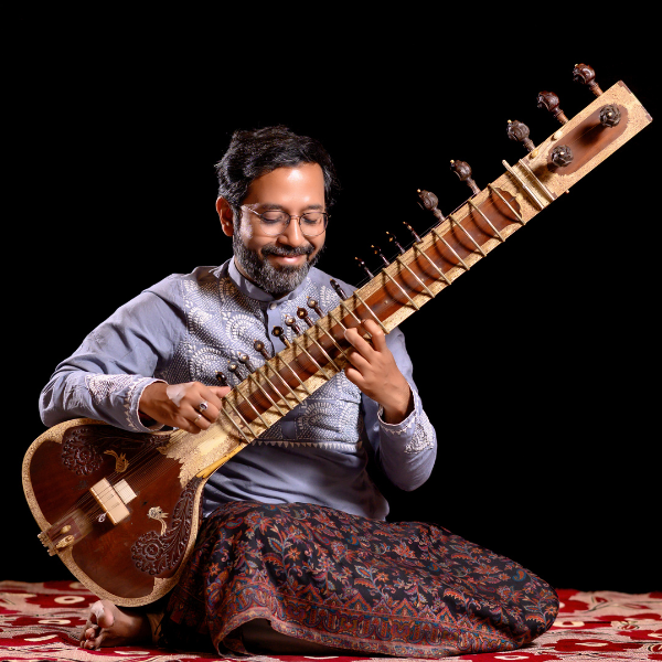 A musician sits cross‑legged on a patterned red carpet, playing a sitar. They wear a light grey embroidered kurta and patterned trousers, with a black backdrop behind them.